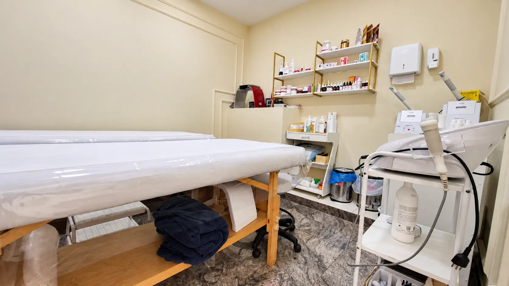 Sala de procedimentos com macas cobertas e prateleiras douradas com cosméticos